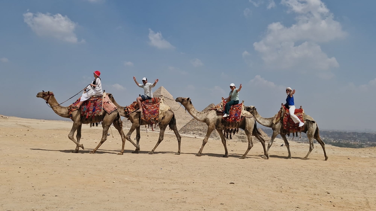 Groupe de touristes à dos de chameaux avec les pyramides au loin.