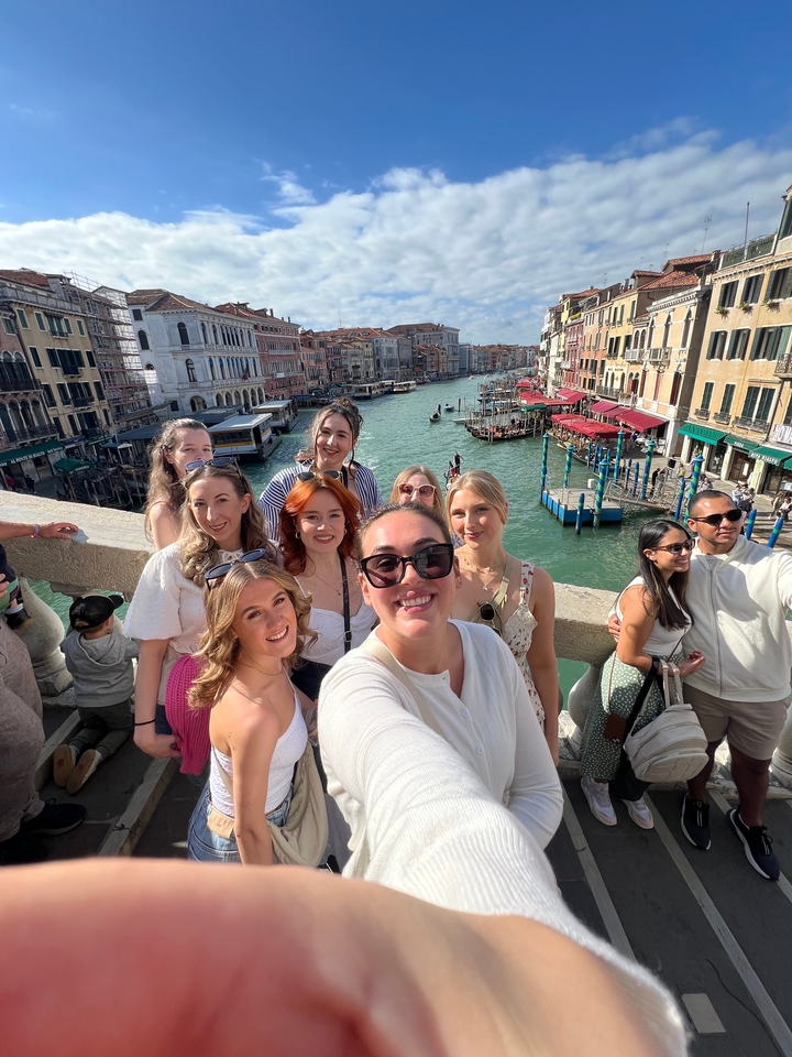 Groupe d'amis posant sur un pont avec un canal et des bâtiments historiques en arrière-plan.