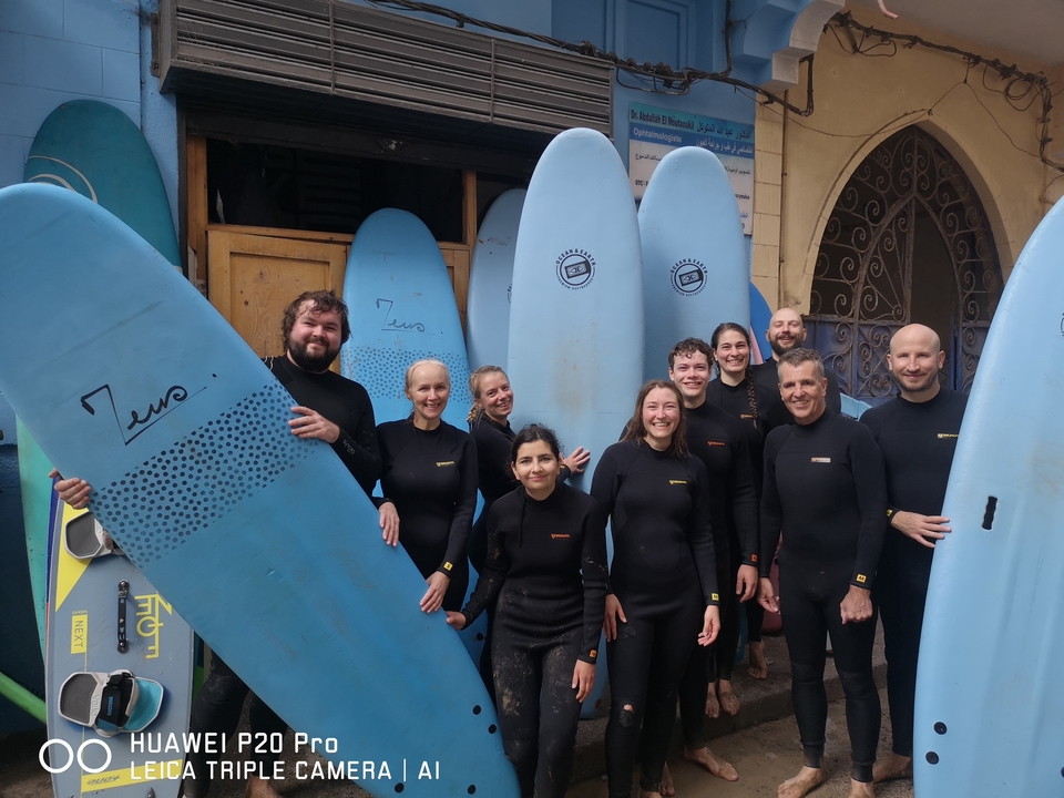 Menschen, die mit Surfbrettern posieren und Neoprenanzüge tragen, stehen vor einem Gebäude.