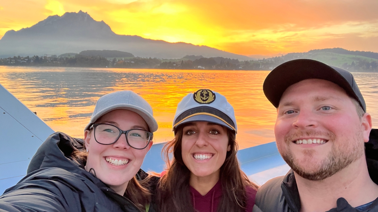 Trois personnes souriantes avec un magnifique coucher de soleil sur un lac et des montagnes en arrière-plan.