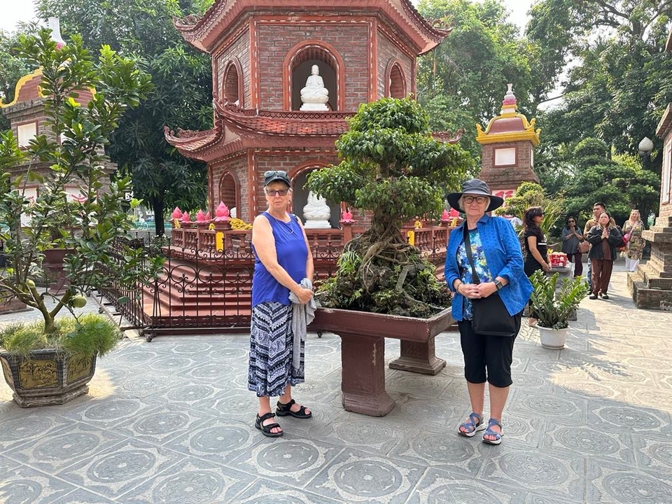 Travellers stand beside a bonsai tree in a colourful courtyard of an ornate pagoda.