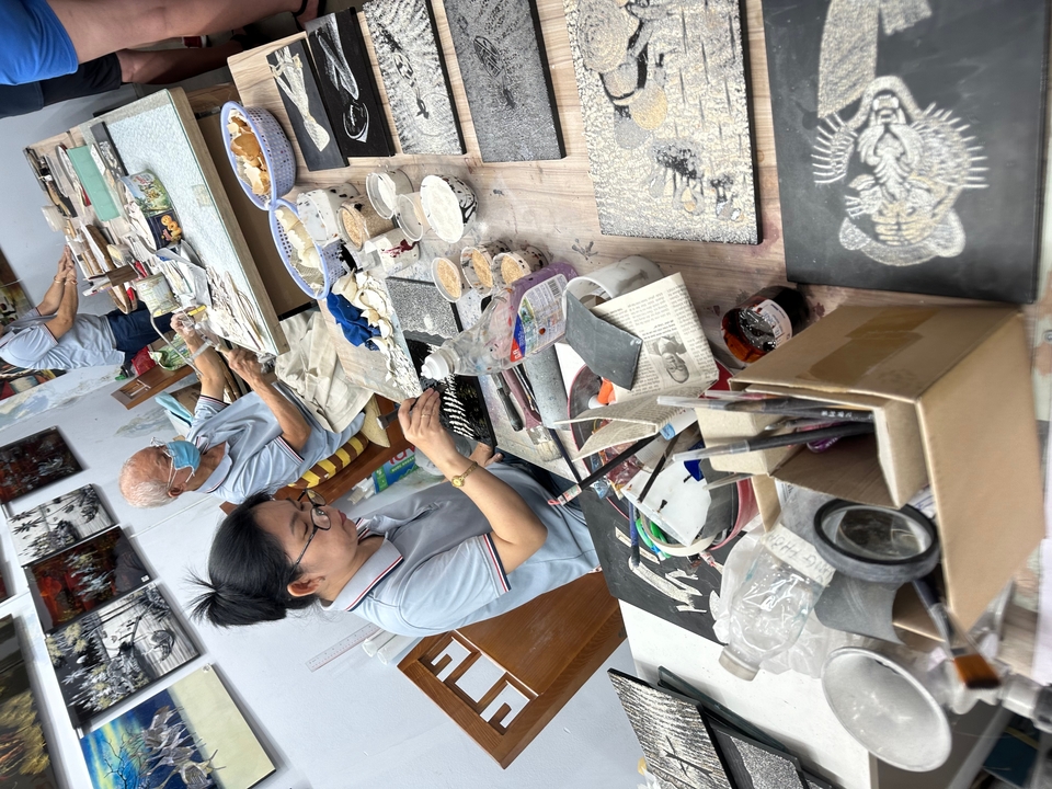 Artisans work on intricate lacquerware pieces at a crowded workshop table covered in tools and materials.