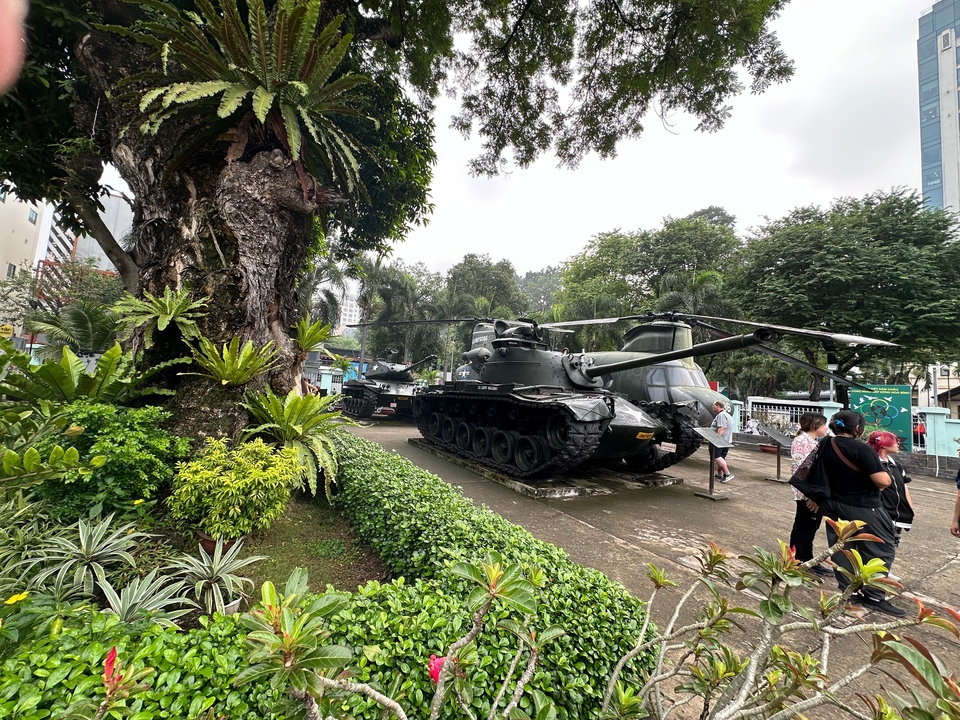 Military tanks displayed in a lush outdoor museum setting while visitors walk between exhibits.
