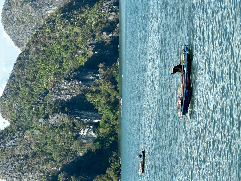 Long wooden boat gliding across turquoise water with towering limestone cliffs covered in greenery behind it.