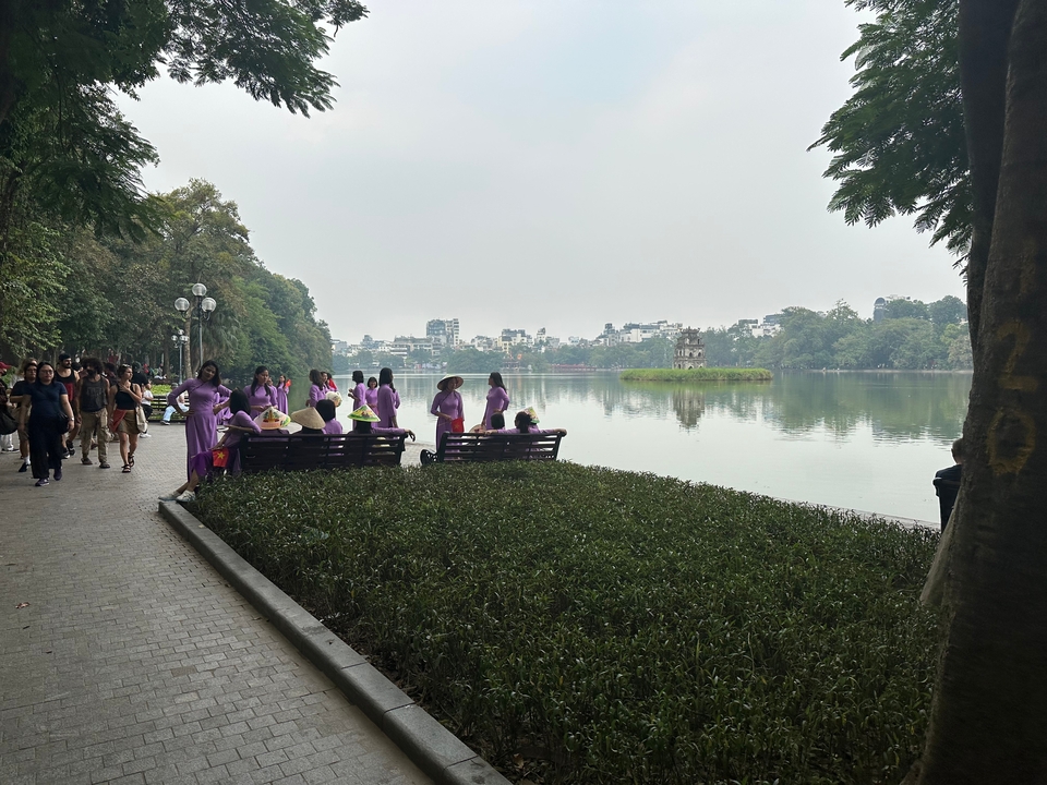 Local women in purple traditional dress gather on benches beside a calm lake with a small tower on an island and city skyline in the distance.