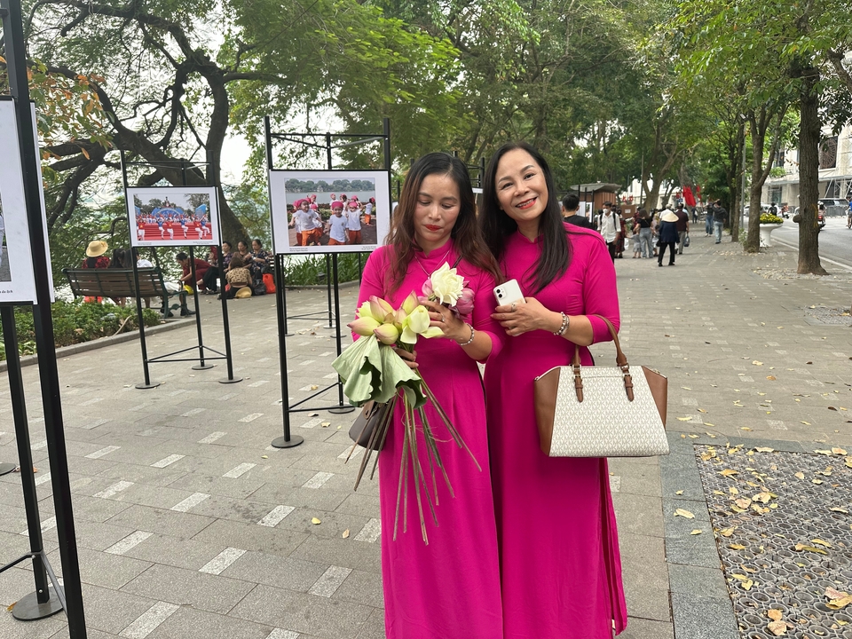 Two smiling women in bright pink ao dai holding lotus flowers on a tree-lined sidewalk gallery.