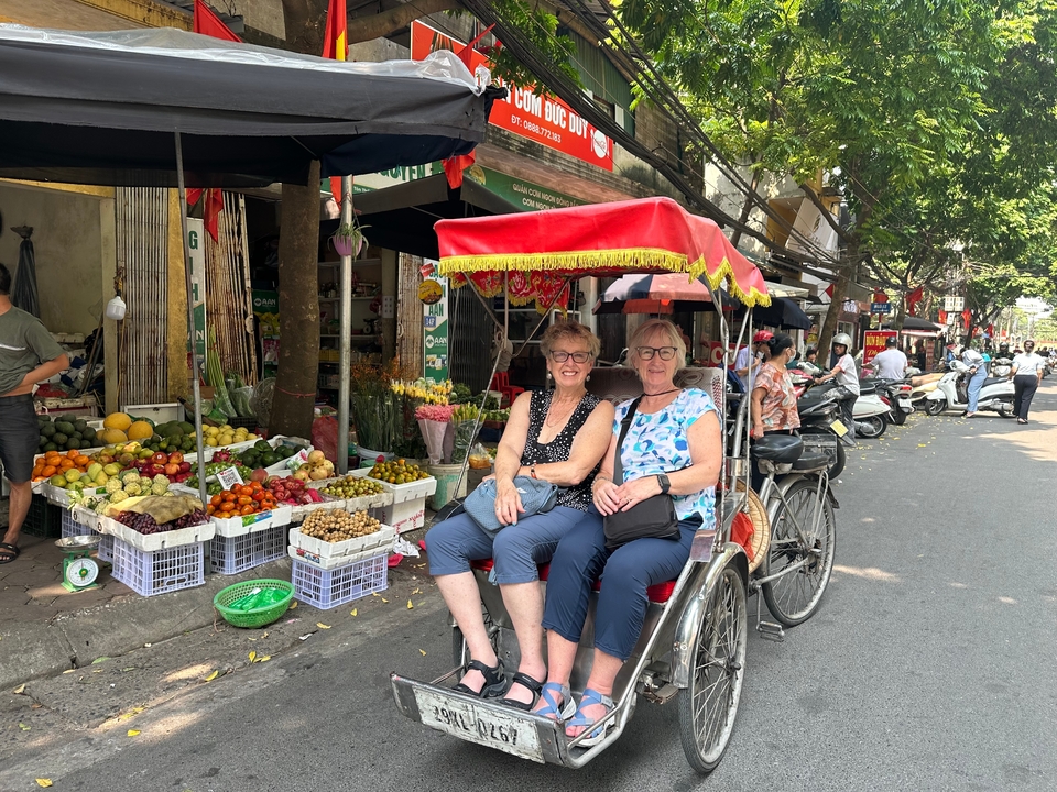 Two tourists seated in a colorful cycle-rickshaw on a busy Vietnamese market street lined with fruit stalls.