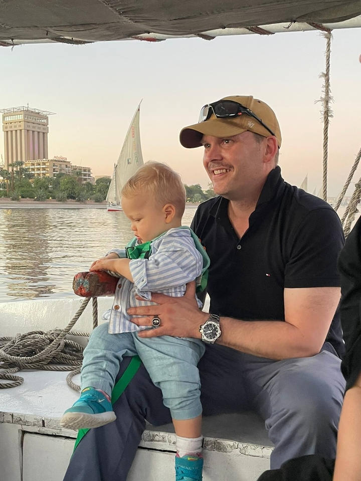 Un homme tenant un enfant sur un bateau avec une rivière et des bâtiments en arrière-plan.