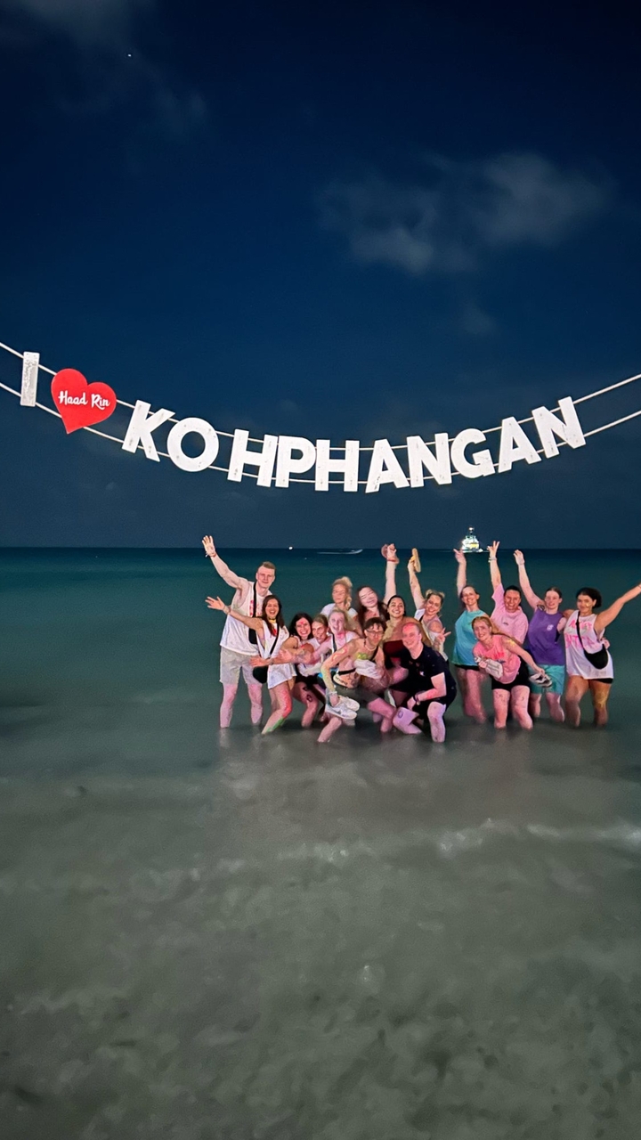 Groupe de personnes debout dans l'eau sous un panneau 'KOH PHANGAN' la nuit.