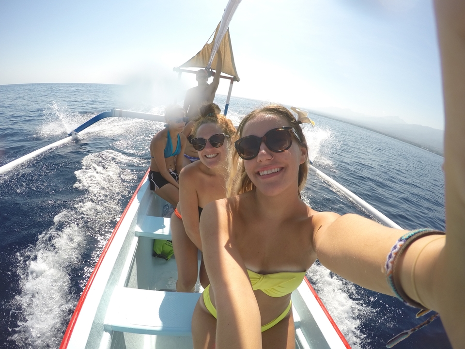 Travellers ride in a small wooden boat across open blue water with spray splashing alongside.