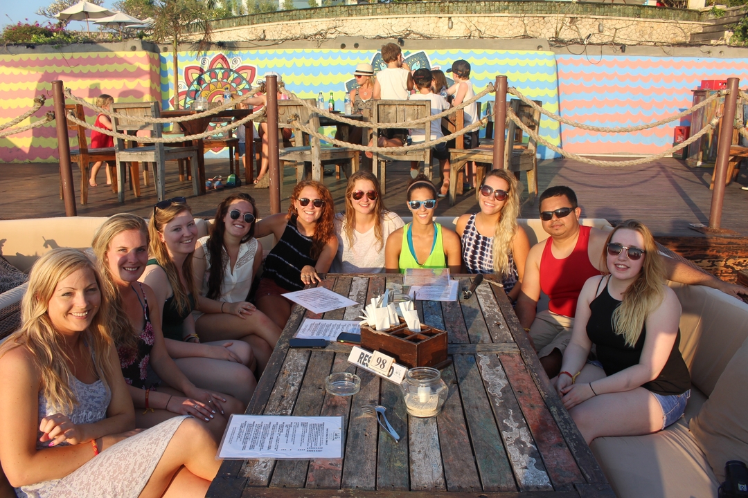 Young travellers sit around a rustic wooden table at a colourful open-air beach bar during golden hour.
