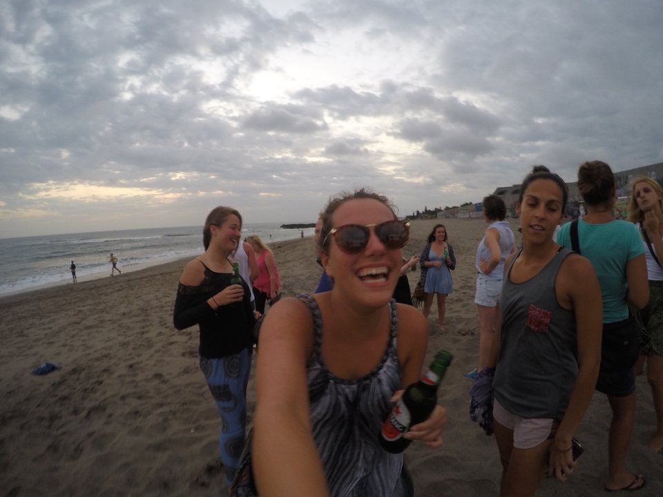 Group of travellers laugh and drink beers on a cloudy beach in the early evening.