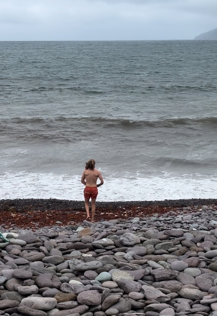 A person standing on a pebble beach looking at rough sea waves.
