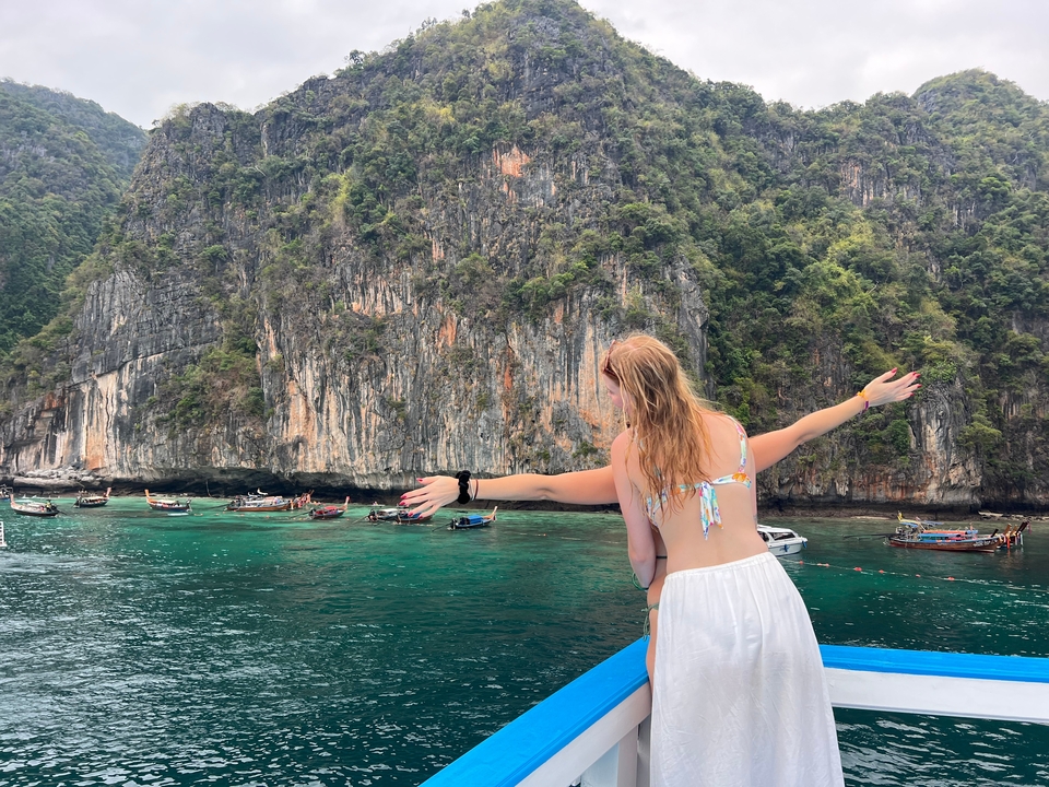 Une personne debout sur un bateau, les bras étendus, avec des falaises et des bateaux en arrière-plan.