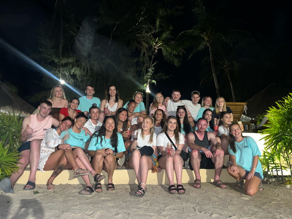 Un grand groupe de personnes posant ensemble la nuit, avec des plantes tropicales et des arbres en arrière-plan.