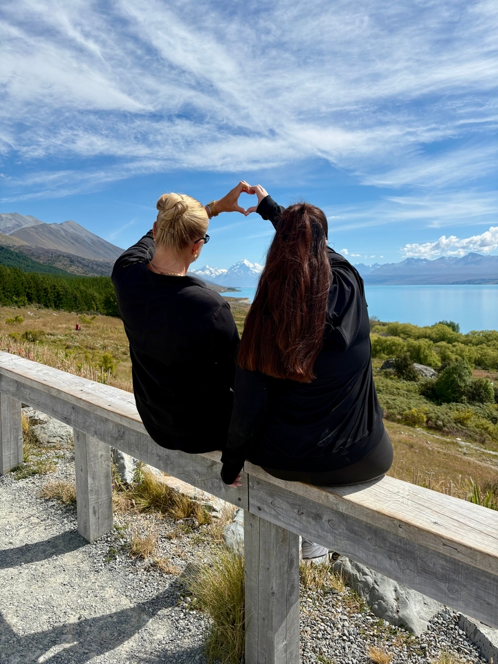 Deux personnes assises sur une rambarde formant un cœur avec leurs mains, surplombant un lac et des montagnes.