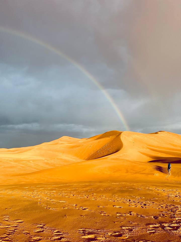 Sanddünen in der Wüste mit einem Regenbogen und stürmischem Himmel.