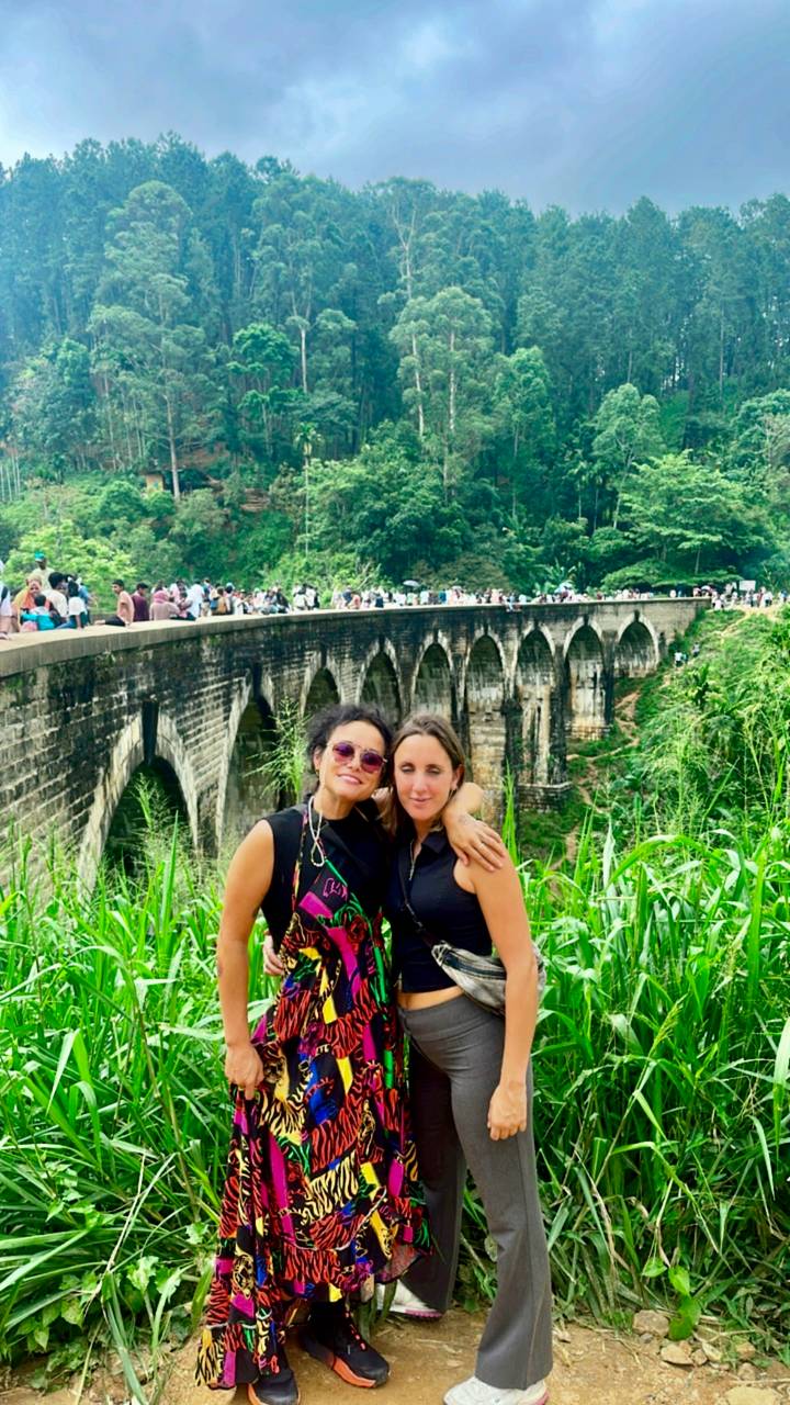 Deux femmes posant devant un pont historique avec une végétation luxuriante.
