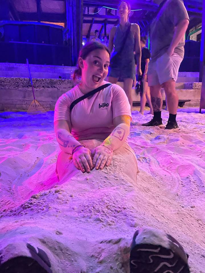 Personne recouverte de sable sur une plage la nuit sous des lumières violettes et roses.