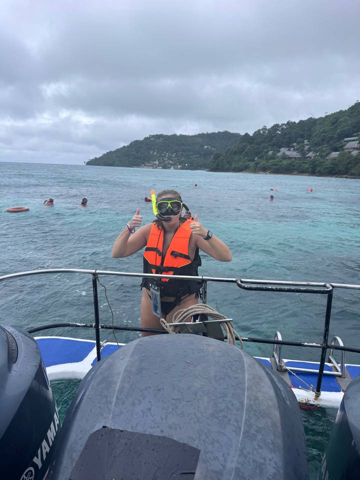 Personne en équipement de plongée avec tuba et gilet de sauvetage sur un bateau, faisant un pouce levé avec l'océan en arrière-plan.