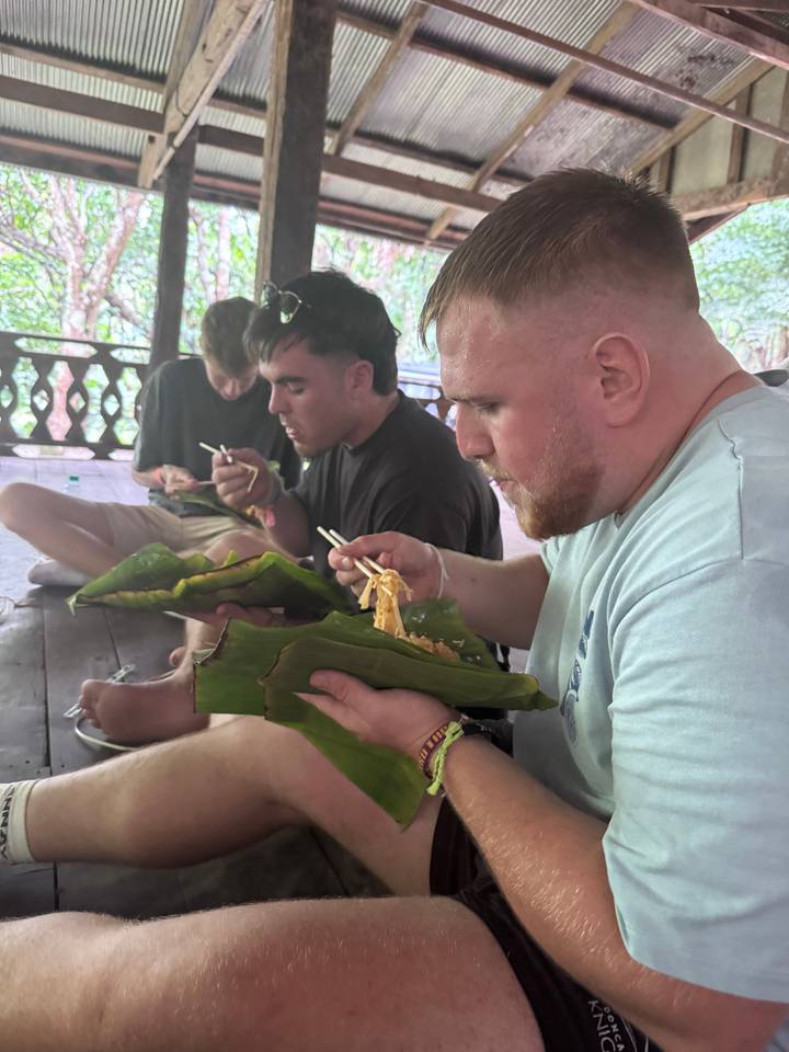 Des gens mangeant de la nourriture enveloppée dans des feuilles, assis sur un plancher en bois.