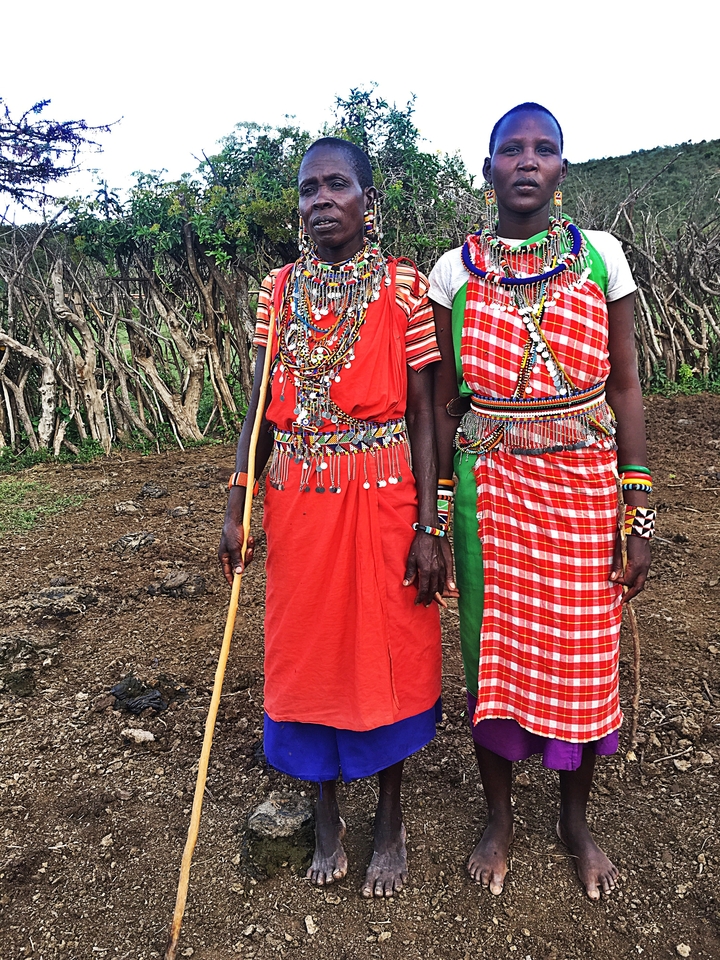Deux personnes en tenue traditionnelle se tenant à l'extérieur, avec du feuillage sec en arrière-plan.
