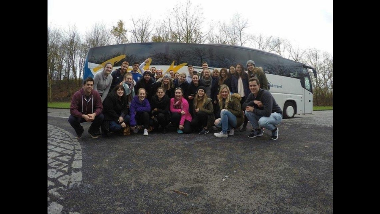A large group poses in front of a travel bus in a parking lot.