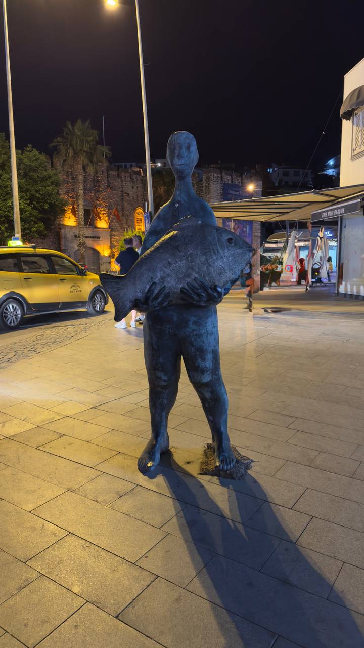 Personne posant avec une sculpture de poisson sur une place la nuit.
