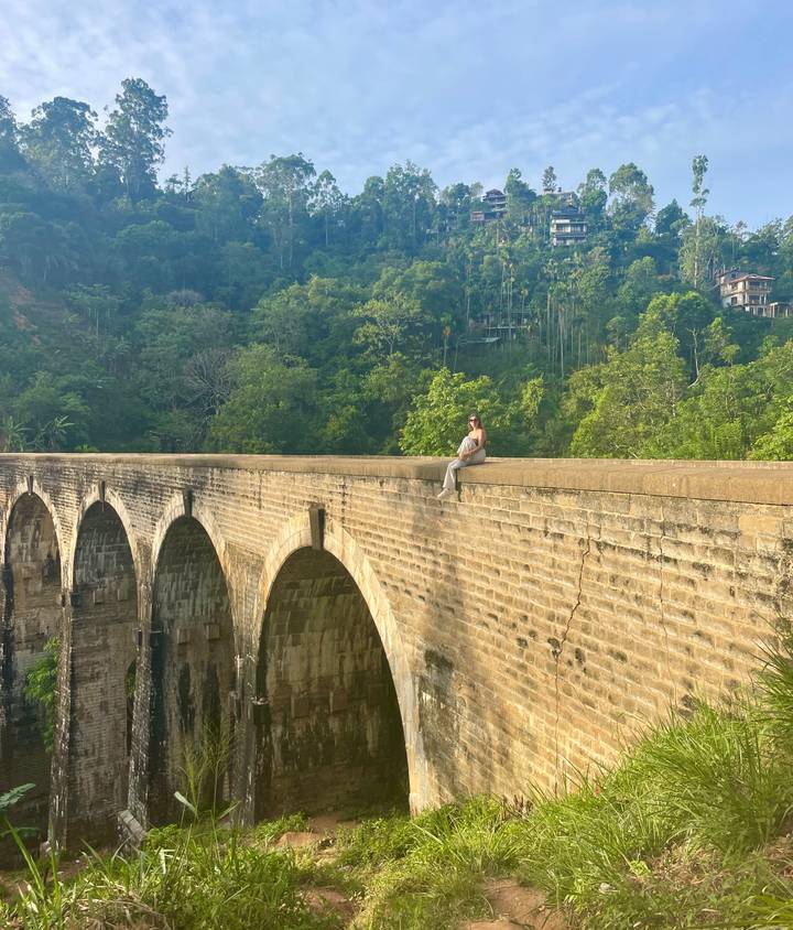 Une personne assise sur un pont historique en arche entourée de verdure.