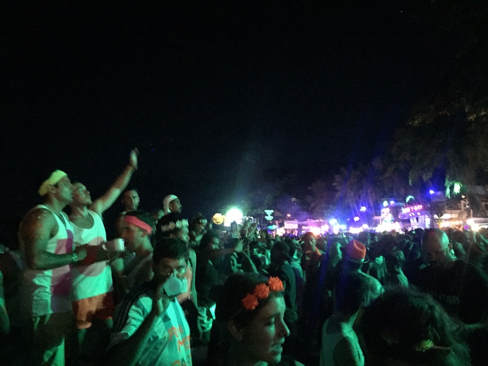 Large crowd dancing at an outdoor night party lit by colorful lights.
