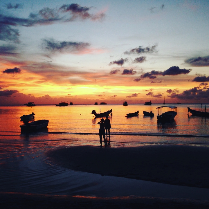 Silhouette of people against a vibrant sunset at the beach with boats on the water.