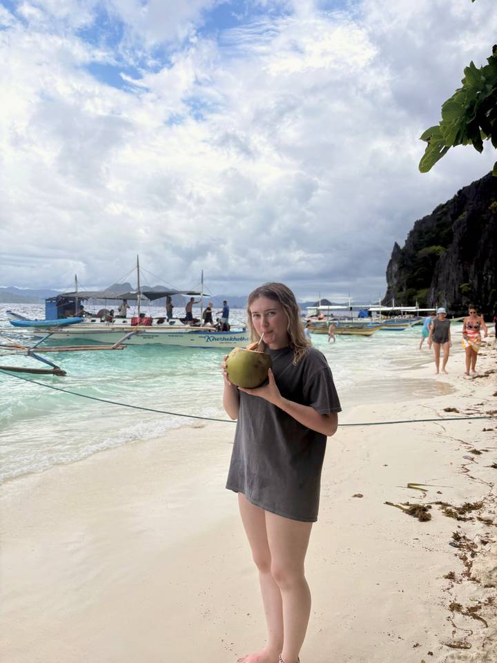 A woman drinking from a coconut on a beach with traditional boats in the background.