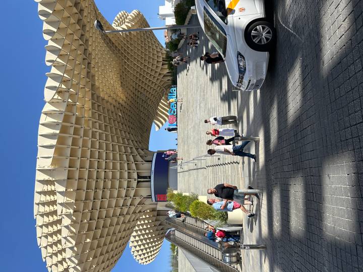 Personnes marchant sous une structure architecturale moderne avec un panneau 'Sevilla'.