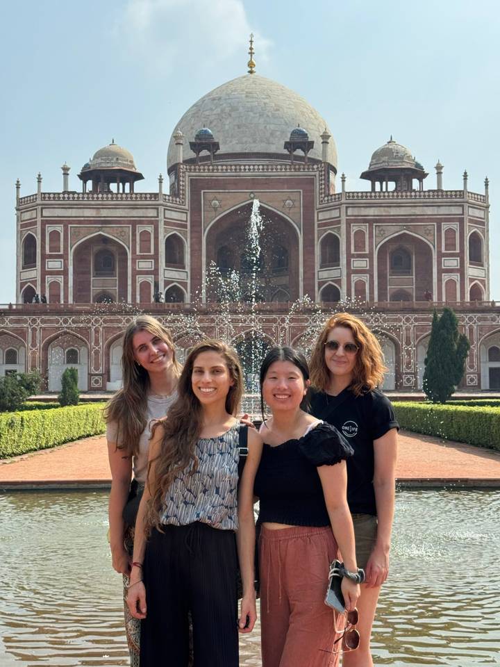 Quatre femmes souriantes devant un bâtiment historique avec des fontaines.