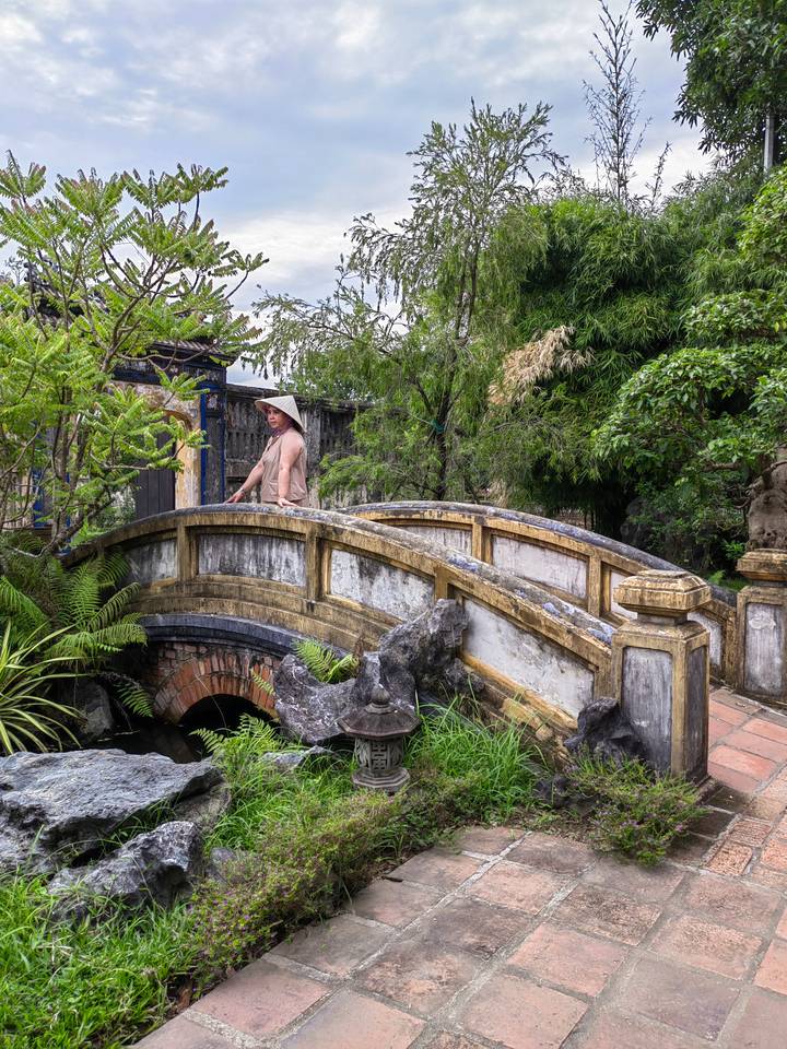 Personne portant un chapeau conique debout sur un pont de pierre pittoresque entouré d'arbres.