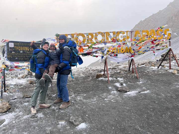 Three people with backpacks at a mountain pass with prayer flags and signs.