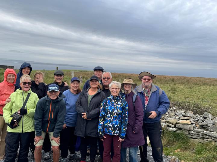 Group photo of smiling tourists in outdoor attire by a stone fence.