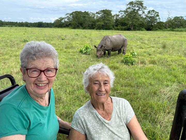Deux femmes souriantes devant un rhinocéros dans un champ herbeux.