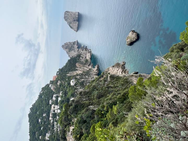 Vue sur falaise avec mer bleue et formations rocheuses, maisons perchées sur des collines verdoyantes.