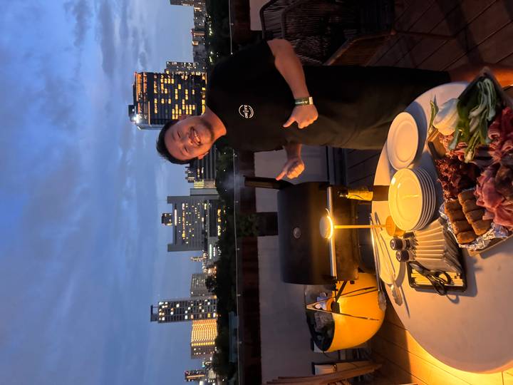 Persona de pie junto a una barbacoa en una azotea con edificios de la ciudad al fondo.