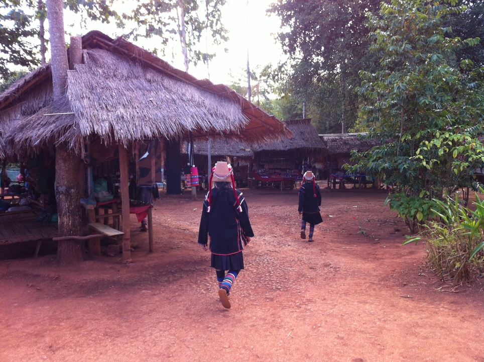 Two hill-tribe women in traditional dark outfits and colourful socks walk through a thatched village market.