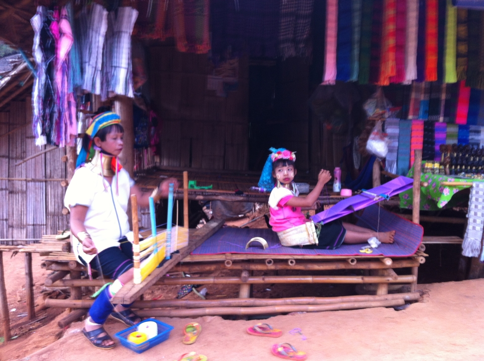 Long-neck Karen artisans demonstrate traditional weaving on wooden looms in a village stall.