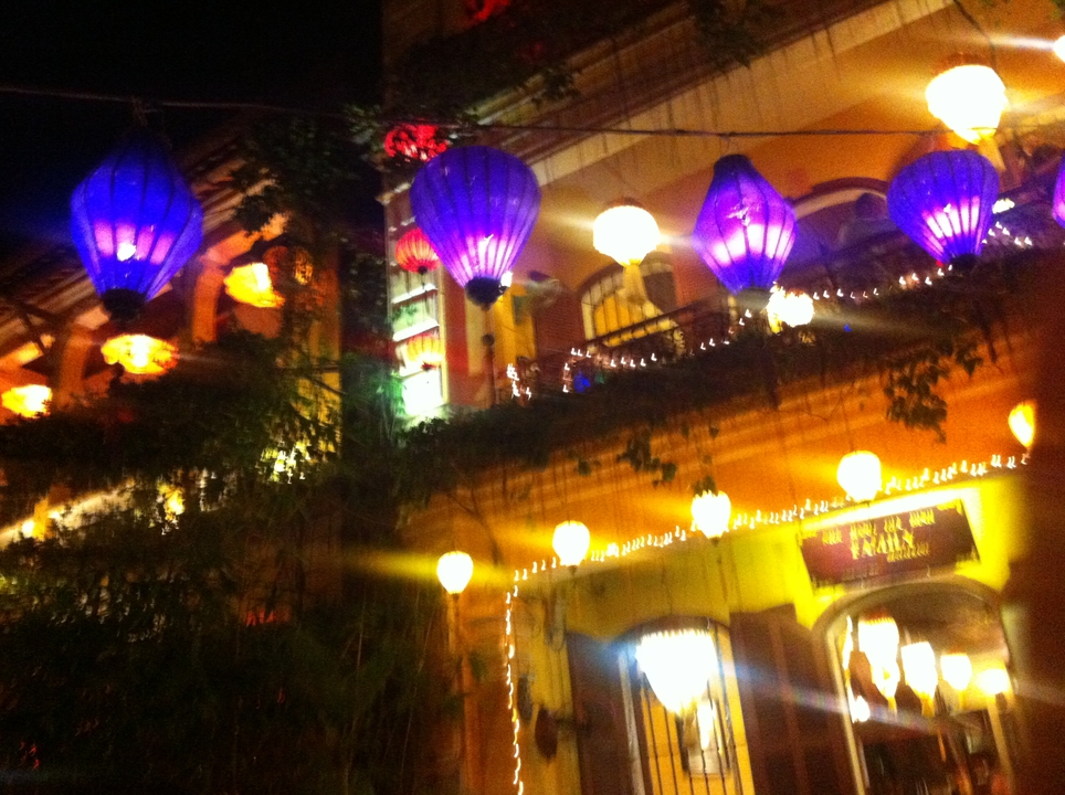Colourful lanterns blur in front of a yellow colonial building lit up at night.