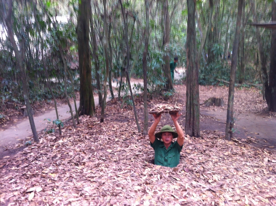 Guide emerges from a camouflaged trap door in the ground at the Cu Chi tunnels, holding leaves overhead.
