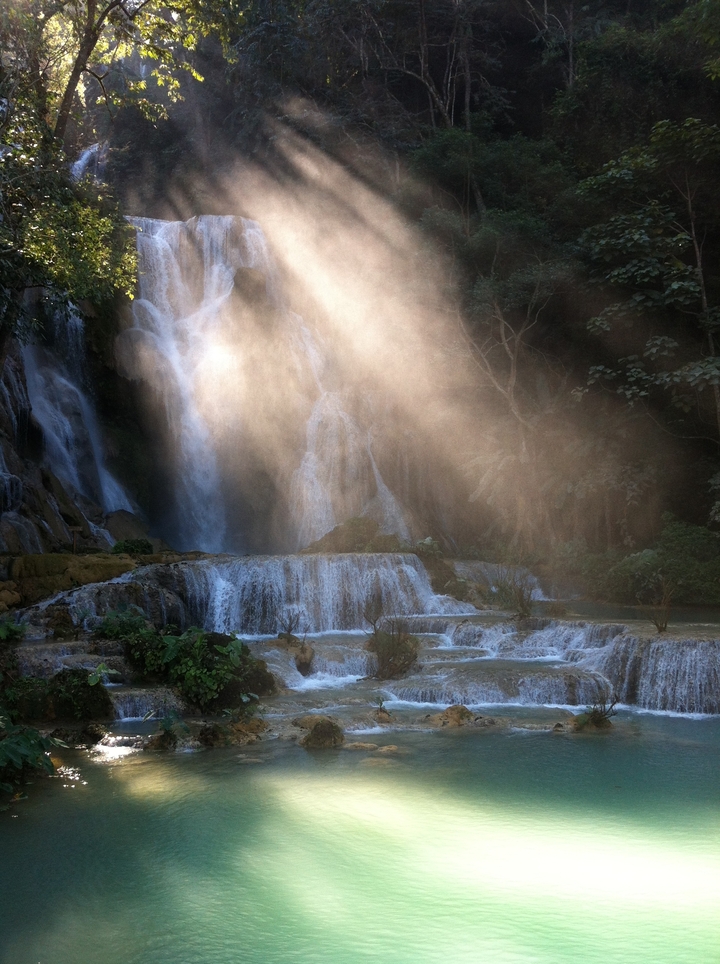 Sunbeam cuts through mist over cascading turquoise waterfall pools in dense forest.
