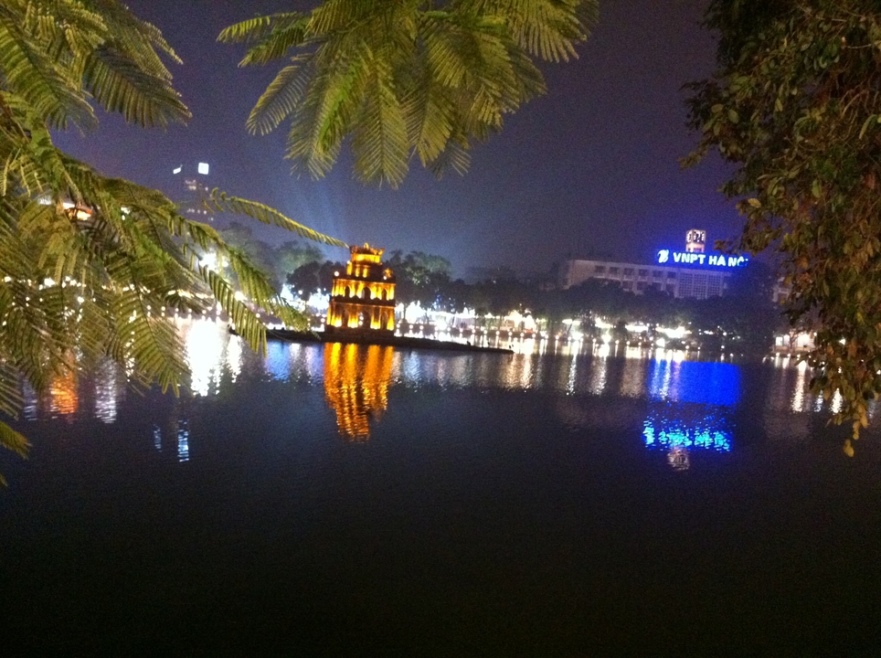 Night view of illuminated Turtle Tower reflecting in Hanoi’s Hoan Kiem Lake with city lights.