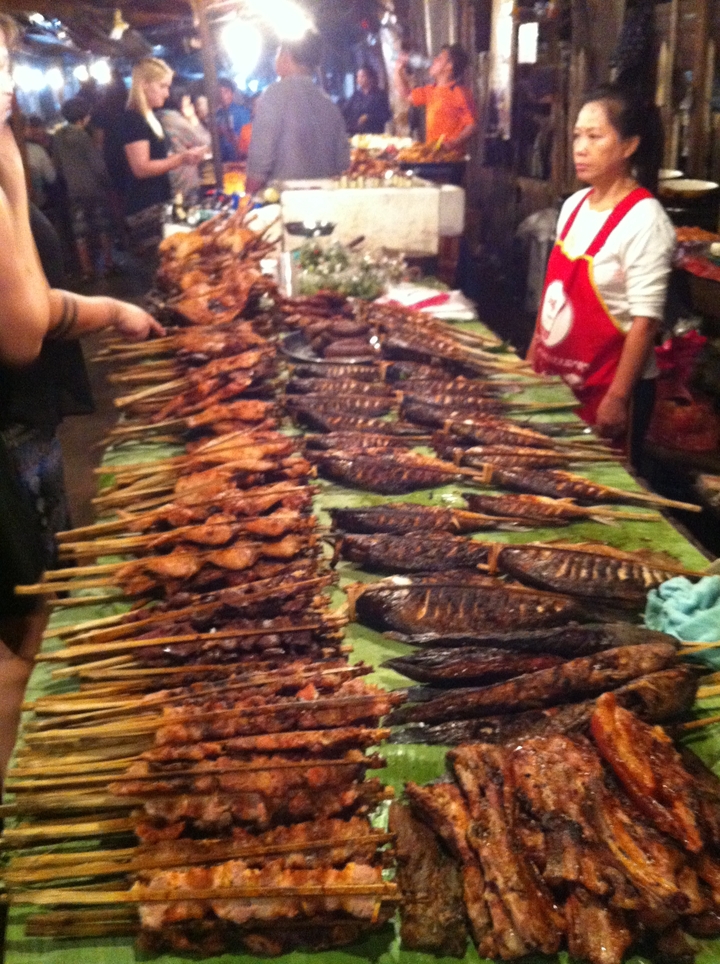Long rows of grilled fish and meats on banana leaves at a bustling night food market.