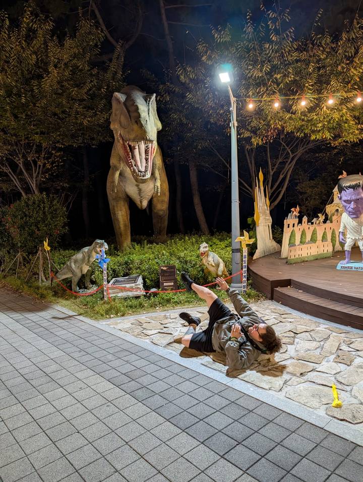 Persoon poseert speels met dinosaurusbeelden in een park tijdens de nacht.