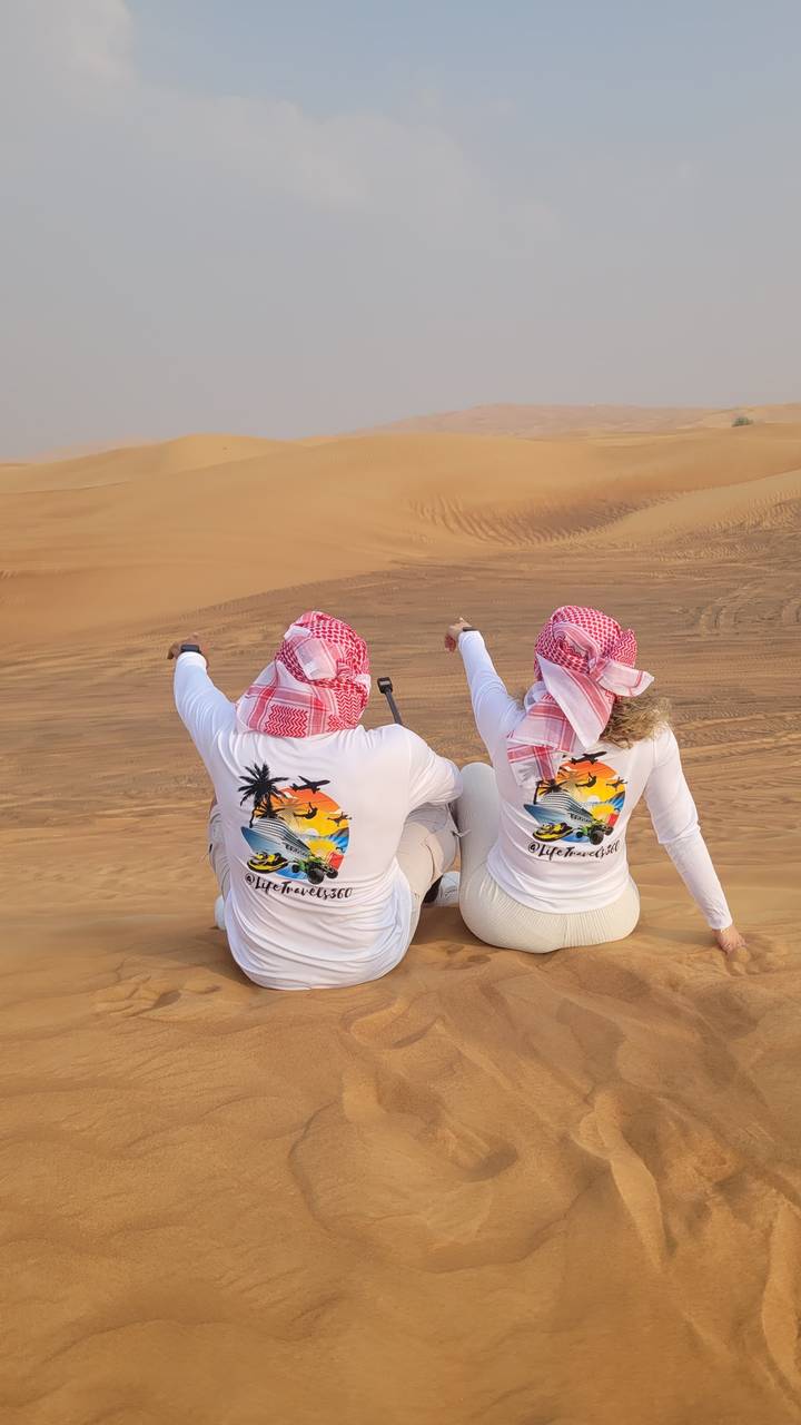 Zwei Personen sitzen auf Sanddünen, tragen passende Outfits und blicken zum Horizont.