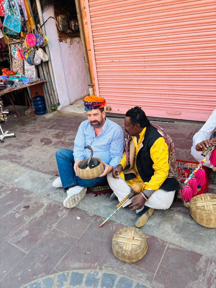 Dos hombres sentados en el suelo, uno usando un turbante tradicional y sosteniendo una serpiente en una canasta.